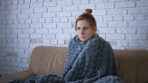 Covered with grey plaid young caucasian woman freezing feeling cold at home, ill sick girl having fever flu influenza temperature symptoms wrapped in blanket shivering indoors. 