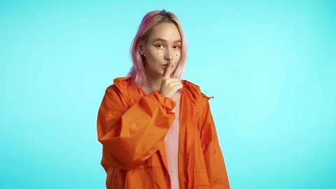 Hipster woman with dyed pink hair holding finger on her lips over blue background studio wall. Gesture of shhh, secret, silence