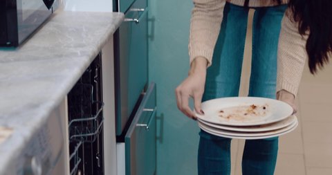 Cinematic view to the mature woman is putting a white dirty plates in the dishwasher. White caucasian female with long brown hair in cozy beige knitted sweater. Cleaning after dinner