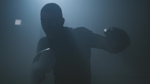 Male boxer punching to camera. Silhouette of young boxer practicing boxing punches looking at camera in dark smoky gym. Close-up in 4K, UHD