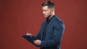 Half-length video of handsome young smiling businessman wearing black shirt and dark blue suit holding the folder taking notes. Dark red background.