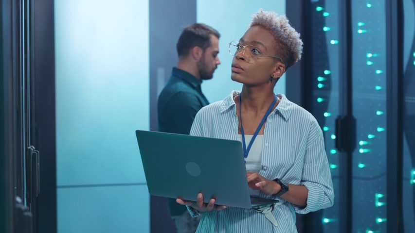 Black female IT engineer upgrading hardware in rack server room corridor working on computer laptop cooperating with male specialist at big data center. Royalty-Free Stock Footage #1041071216