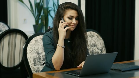 Young modern business woman works on a laptop while sitting in restaurant. Speaking by mobile phone.