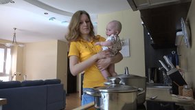Pretty woman in yellow holding baby girl in arms, mix spaghetti meal in kitchen. Woman prepare food in steamy pot. Static shot. 4K UHD video clip.