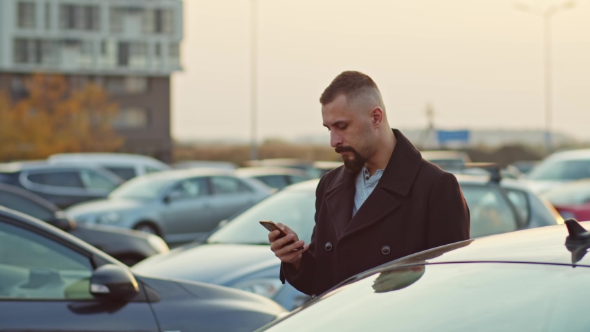 Cheerful bearded man in coat using smartphone social network internet standing outdoor near parked cars. Business person with mobile phone in big city parking space open air at fall.