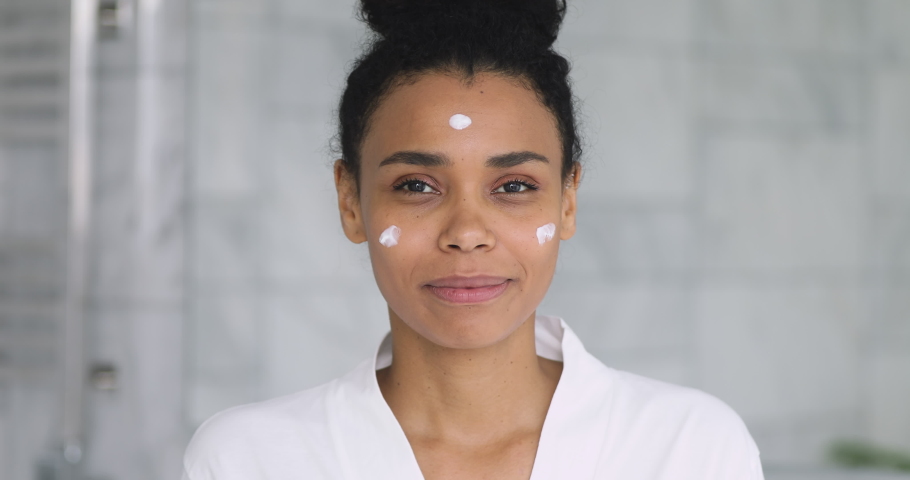Happy beautiful african young woman apply cream on face laugh looking at camera holding creme jar in bathroom pointing aside advertise facial skin care moisture treatment hydration, skincare concept Royalty-Free Stock Footage #1041087274