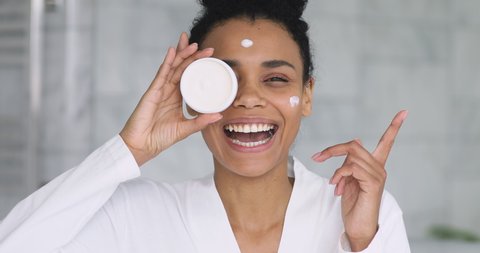 Happy beautiful african young woman apply cream on face laugh looking at camera holding creme jar in bathroom pointing aside advertise facial skin care moisture treatment hydration, skincare concept