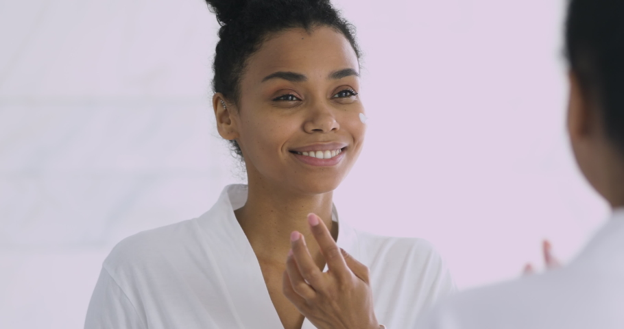 Happy confident attractive young african woman applying cream on face doing morning spa make up routine in bathroom look in mirror enjoy healthy clean soft moisturized hydrated skin, skincare concept | Shutterstock HD Video #1041087277