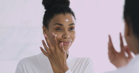 Happy confident attractive young african woman applying cream on face doing morning spa make up routine in bathroom look in mirror enjoy healthy clean soft moisturized hydrated skin, skincare concept