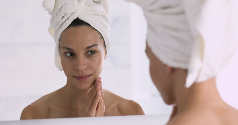 Smiling pretty young woman with towel on head touching face enjoying healthy soft hydrated moisturized skin after removing makeup or applying cream looking in mirror, skincare treatment concept Royalty-Free Stock Footage #1041087310