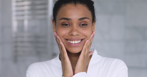 Happy attractive african young woman touching hydrated moisturized healthy facial skin look at camera, smiling mixed race lady enjoying beauty care moisture skincare treatment hydration in bathroom