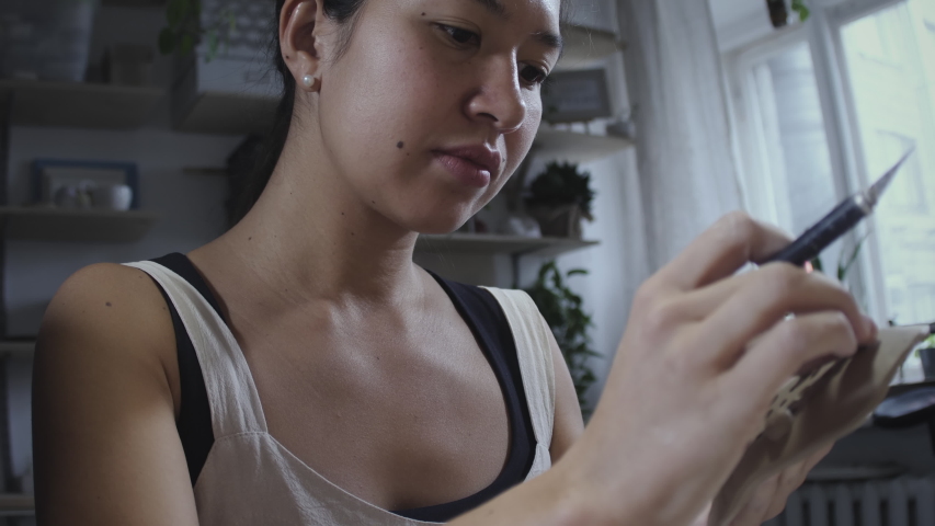 Portrait of young asian master potter holds in her hand soap dish made of raw clay. Potter master works in workshop with knife and molds earthenware