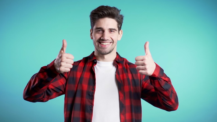 Positive young man smiles to camera. Hipster guy showing thumb up sign over blue background. Winner. Success. Body language.