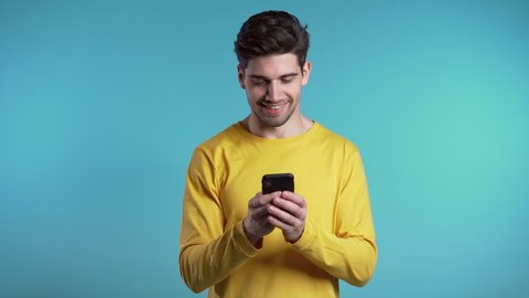 Young man smiling and using mobile phone over blue background. Handsome european guy holding and texting with smart phone. Boy with technology.