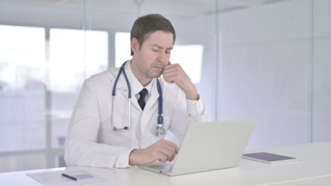 Middle Aged Doctor Thinking and Working on Laptop 