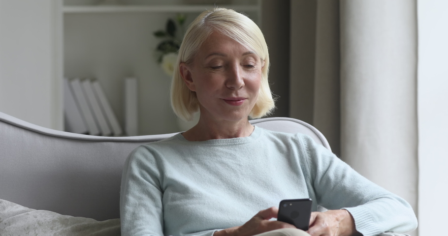 Smiling middle aged older woman enjoying using smartphone sit on couch at home, happy mature adult lady holding handy device texting messages, playing games in mobile app looking at phone screen Royalty-Free Stock Footage #1041108271