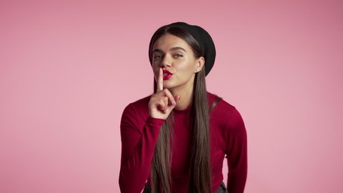 Beautiful woman with red lips holding finger on her lips over pink background. Gesture of shhh, secret, silence. Close up.