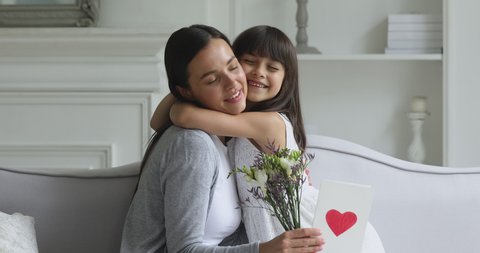 Cute little funny kid child daughter congratulating happy young mom with mothers day concept presenting spring flowers and greeting card embracing affectionate mum having fun sit on sofa at home