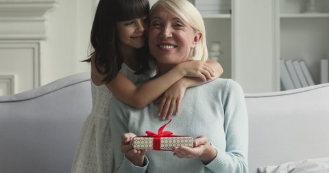 Happy cute little child granddaughter embracing kissing old middle aged grandma foster care parent presenting gift box making surprise congratulating with birthday holiday hugging having fun at home