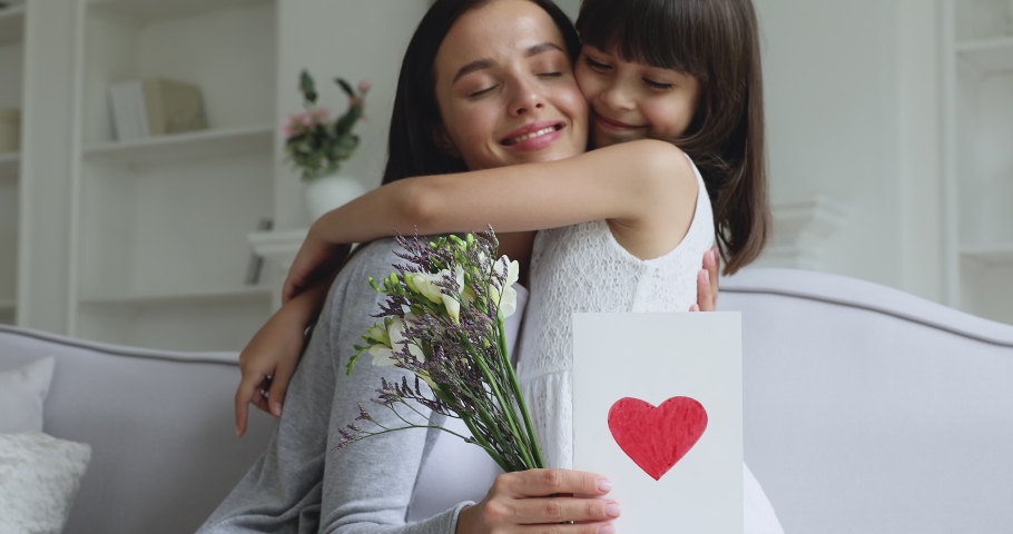 Loving cute small kid child daughter hugging kissing happy young mum congratulating with mothers day concept embracing grateful mommy holding spring flowers and greeting card with red heart at home Royalty-Free Stock Footage #1041120058