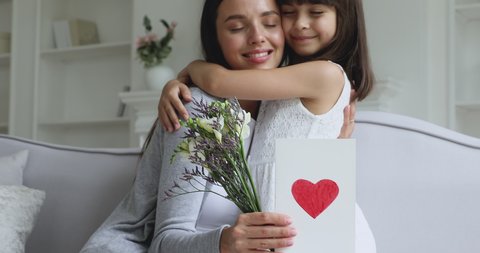 Loving cute small kid child daughter hugging kissing happy young mum congratulating with mothers day concept embracing grateful mommy holding spring flowers and greeting card with red heart at home