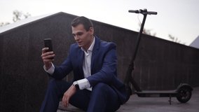 Attractive businessman sitting on the stairs and does video cahtting on smartphone outside. Man in elegant blue suit having video call with collegues. His scooter behind him. Slow motion