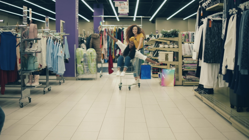 Cheerful lady is wheeling her female friend in a shopping trolley