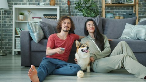 Happy couple girl and guy are watching TV smiling and hugging corgi doggy at home, man is holding remote control. Youth, family and entertainment concept.