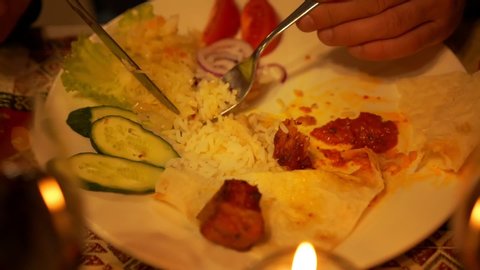 woman puts crumbly rice on fork with knife at cucumber slices and red sauce on plate among candles closeup slow motion