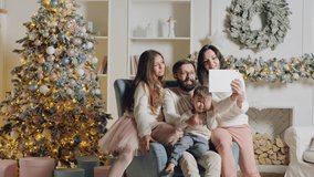Happy young family with two children sitting on the armchair in the living room , online video chatting. The room is decorated with a Christmas tree and garlands. Everyone waving their hands while