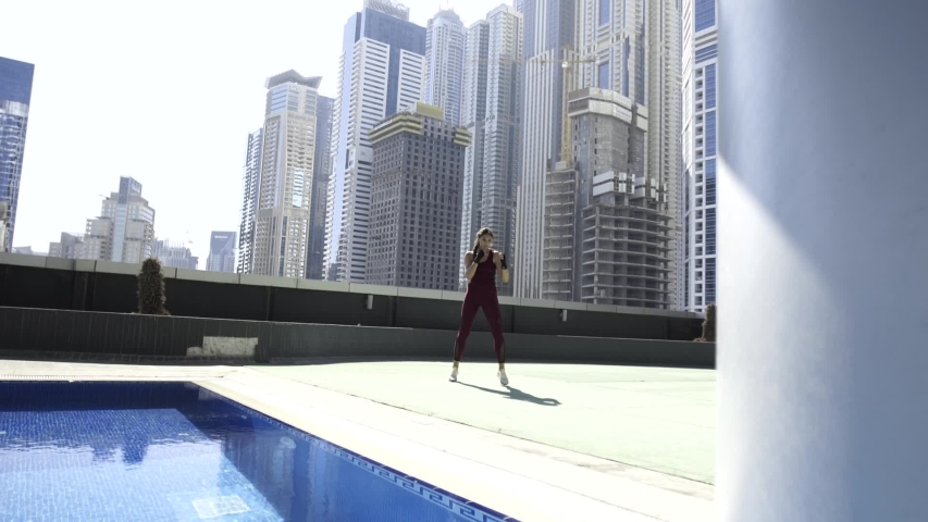 Beautiful young leggy kickboxer girl is training on the roof of the building along the pool doing high kicks and punches. City view of skyscrapers. Wide shot. Slow motion.