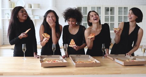 Happy cheerful funny attractive diverse young women best friends wear elegant black dresses drink champagne laugh share eat takeaway pizza together in kitchen celebrate hen sorority party together