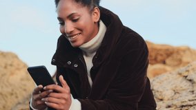 Beautiful cheerful African American girl in down jacket laughing happily using cellphone on stones by the sea