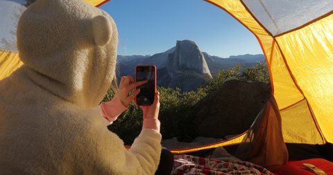 A girl in a tourist tent wearing a funny sweater with the ears of a teddy bear takes the phone picture of the famous Half Dome rock, located in the eastern part of Yosemite Valley in California, 2019