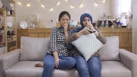 Two frightened islam and japanese female friends watching thriller movie at home kitchen. malay girl holding pillow and tv control. Multiracial young women scared by horror film on television.