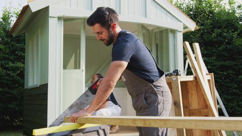 Carpenter sawing wood in foreground as he and apprentice build summerhouse in garden - shot in slow motion