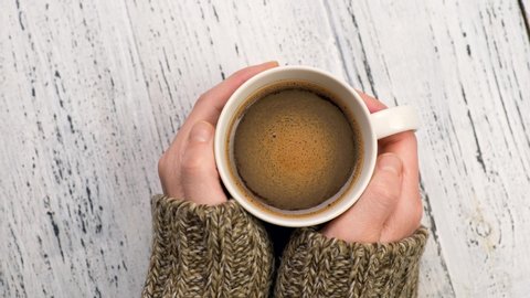 Woman drinking coffee and warms her hands. Female hands in sweater holding cup of morning coffee. Shabby white rustic background 스톡 비디오