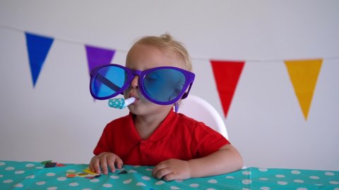 Cute boy with huge glasses and party noisemaker have fun by birthday afterparty table. Confetti shot. Gimbal motion shot.