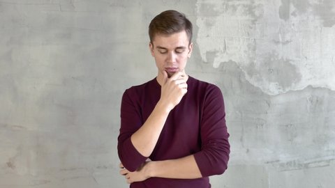 Portrait of a young attractive man in a burgundy jumper stands against a gray uneven wall, pokes his chin with his right hand and finds a solution to the problem raises his finger