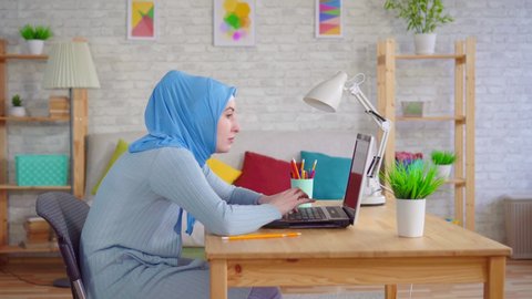 young Muslim woman in a traditional headscarf slouches while sitting at a laptop and feels pain in her neck