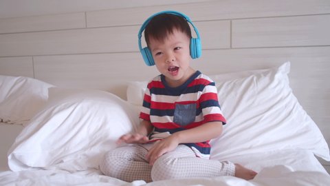 Cute smiling Asian 3 - 4 year old toddler boy child listening to music in headphones & singing in bed. Little kid listening loud music in wireless earphones and sing along and dancing at home 
