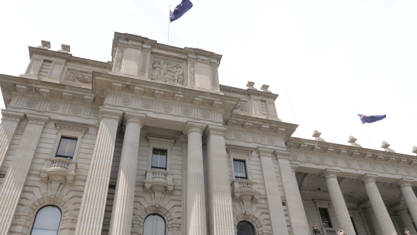 View of the facade of the Parliament House of the state of Victoria in Melbourne, Australia. Flags of Australia and Victoria state waving on the top, blue sky during a day of spring.