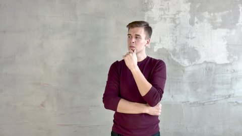 A young man in burgundy jumper short haircut stands against gray wall thoughtfully rubs his chin then laughs waves his hands medium shot
