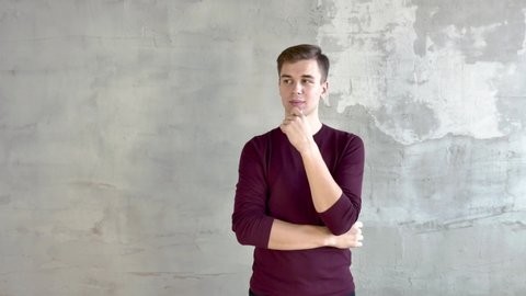 A young man short-cut in a burgundy sweater stands against a gray wall, thoughtfully rubs his chin, then laughs, waves his hands. The average plan.