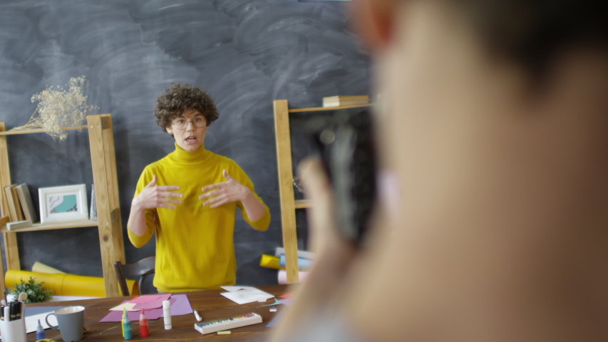 Handheld medium shot of young curly haired female decorator in eyeglasses and yellow turtleneck talking about art supplies while cameraman recording video blog for her