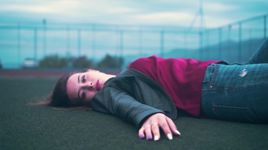 A beautiful and passionate girl in a biker jacket and jeans lies on the floor of a basketball or tennis court. A young woman is lying on the surface of a court. The camera moves away from the girl