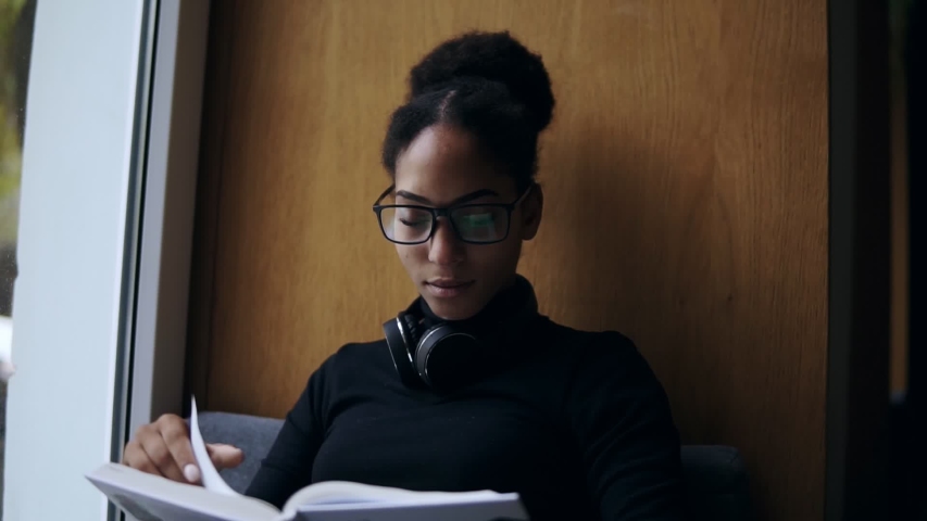 The young attractive afro-american woman is reading the book on the wide window-sill. Close-up portrait of a stylish girl in eyeglasses and headphones reading or studying. Close up | Shutterstock HD Video #1041220342