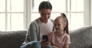 Happy family mom and cute little kid child daughter having fun using smart phone enjoying playing mobile games watching funny videos looking at cellphone screen bonding sitting on sofa at home