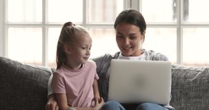 Happy young mom teach little child daughter learn using laptop tech at home, adult mother having fun watch cartoon with kid girl surfing social media online shopping talk look at computer sit on sofa