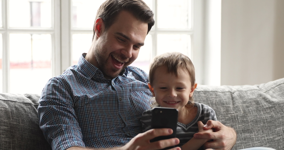 Happy young adult parent dad teaching cute preschool child kid son learning using smartphone apps playing mobile games having fun with cell phone looking at cellphone sitting on couch at home Royalty-Free Stock Footage #1041220366